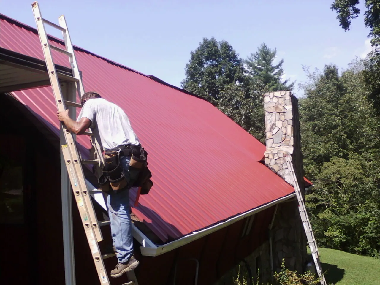 Roofer on a ladder beside a red metal roof and stone chimney — Soffit & Fascia Repair by Roofing Brothers in North Palm Springs